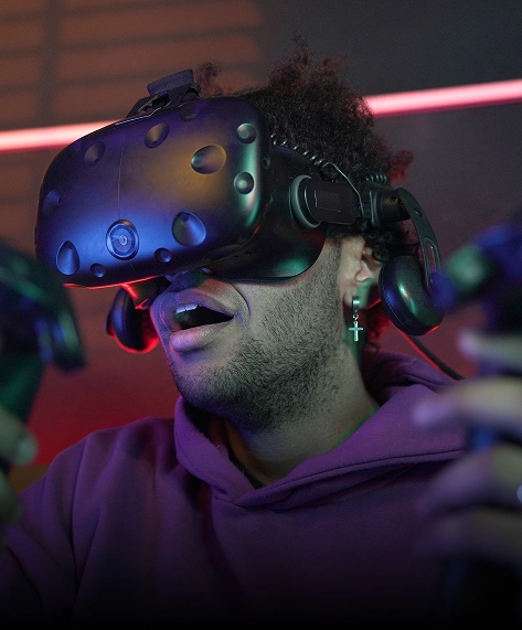 Een persoon met een virtual reality-headset houdt twee controllers vast en lijkt geboeid en verbaasd. Hij draagt een paarse hoodie, met kleurrijke verlichting op de achtergrond